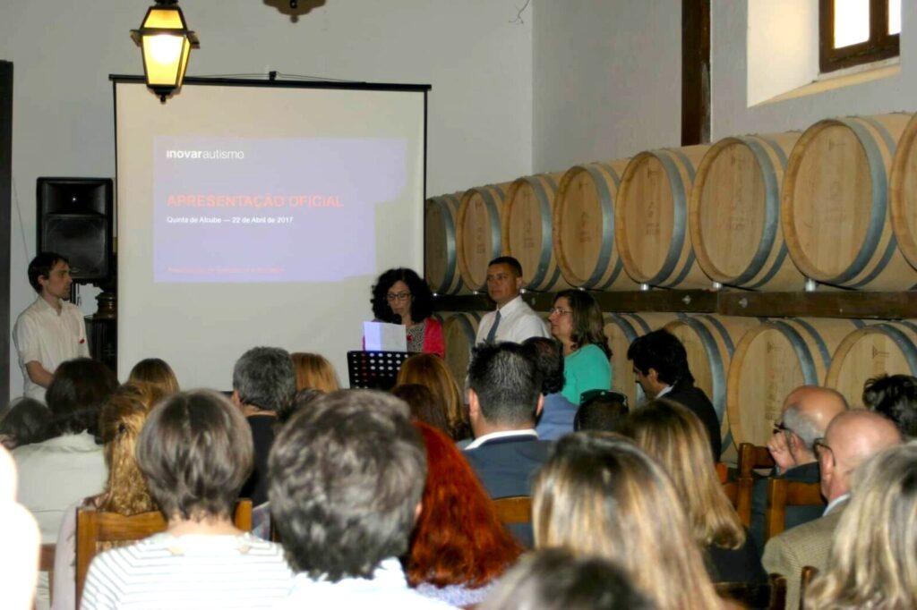 Evento formal realizado em uma sala com paredes revestidas por grandes barris de madeira, sugerindo um ambiente de adega ou vinícola. Diversas pessoas estão sentadas, voltadas para um púlpito onde um homem e duas mulheres fazem uma apresentação.